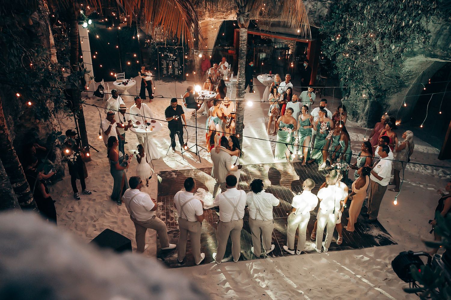 Wedding reception celebration at dusk with guests gathered under tropical foliage.