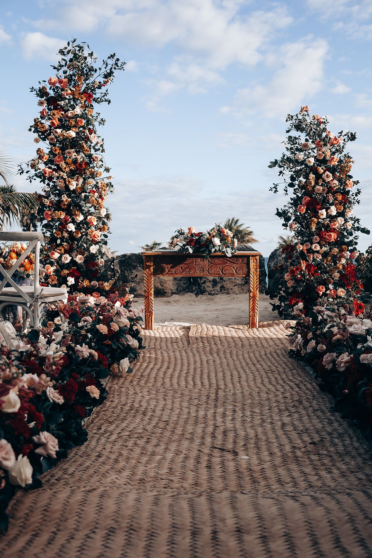 A rustic wooden arch adorned with coral roses lines a cobblestone path under a bright blue sky.