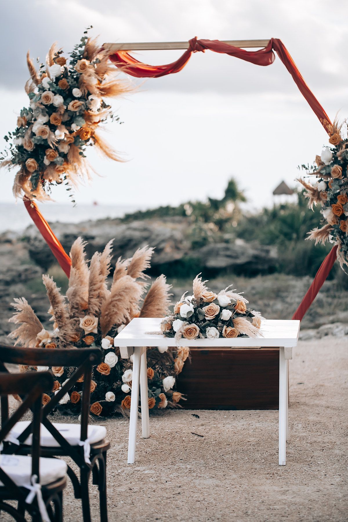 Rustic outdoor wedding ceremony setup with red wooden arch and white benches decorated with dried pampas grass and flowers.