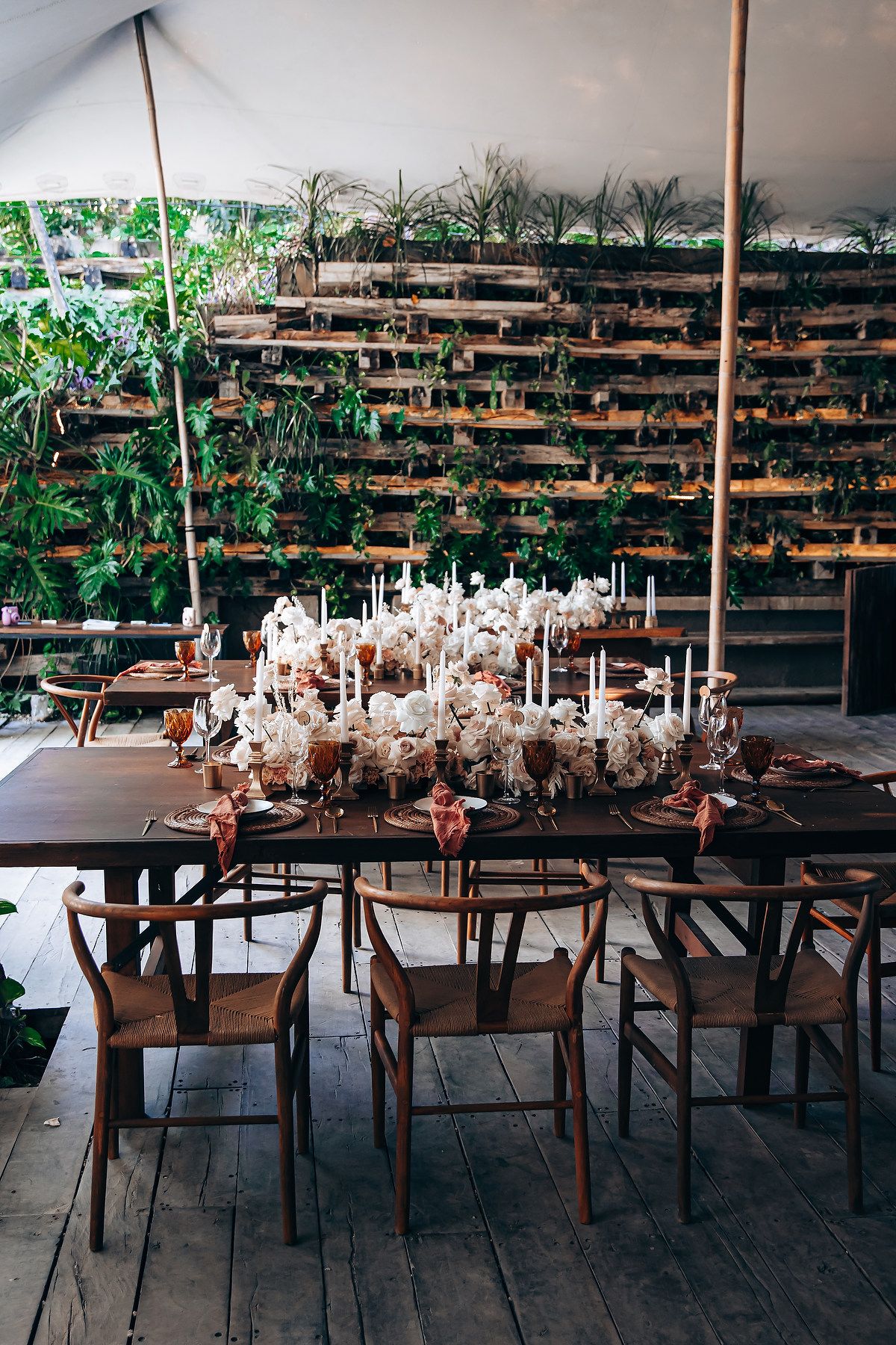 Long dining tables set with white candles and wooden chairs against a backdrop of green plants.