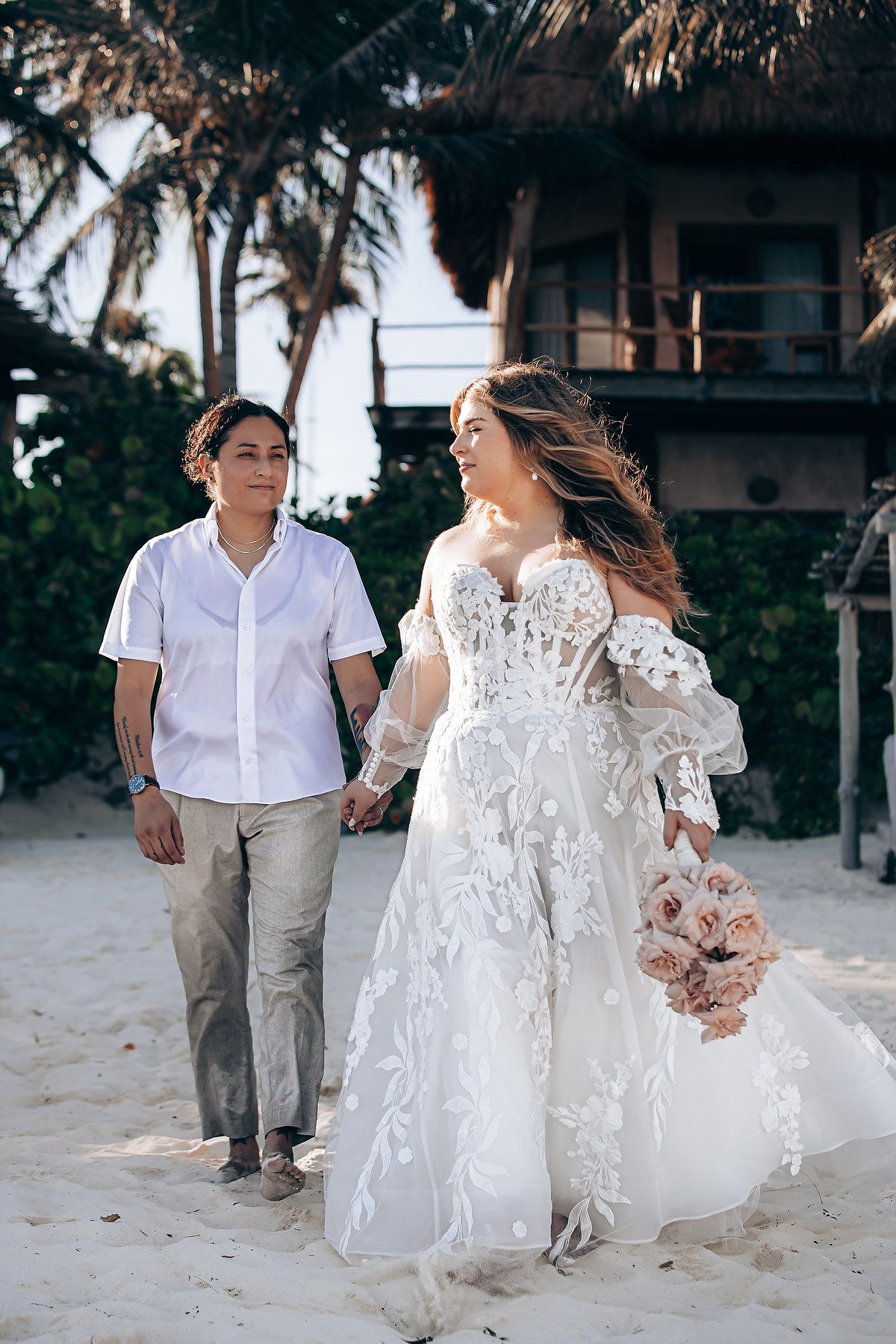 A couple walks along a tropical beach with palm trees and a rustic building in the background.