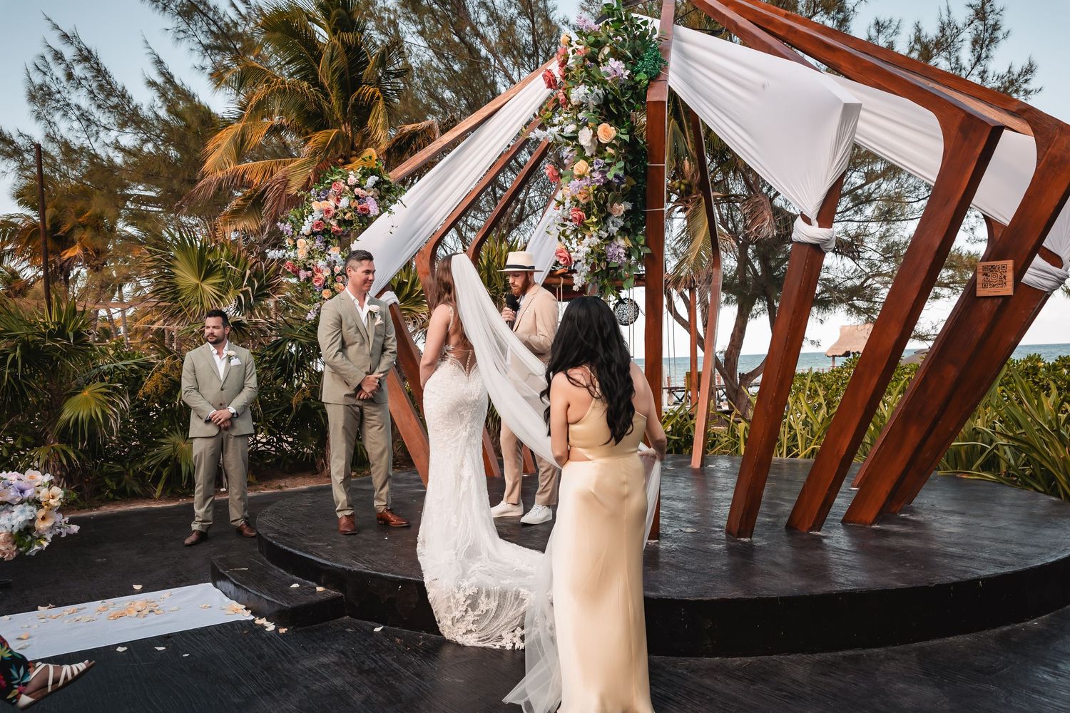 Wedding guests seated in white chairs at tropical beachside ceremony with wooden arch structure.