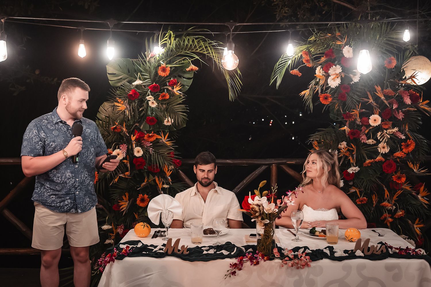 Wedding reception head table decorated with floral arrangements and string lights.