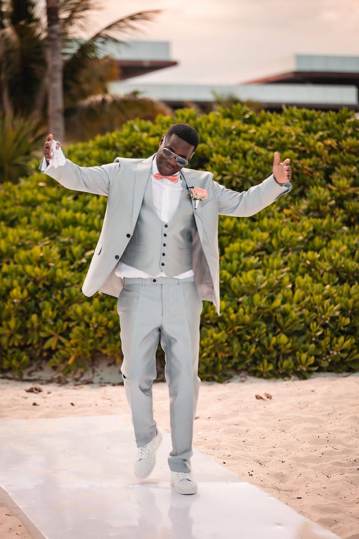 Person in light gray suit dancing joyfully on a sandy beach with tropical foliage in background.