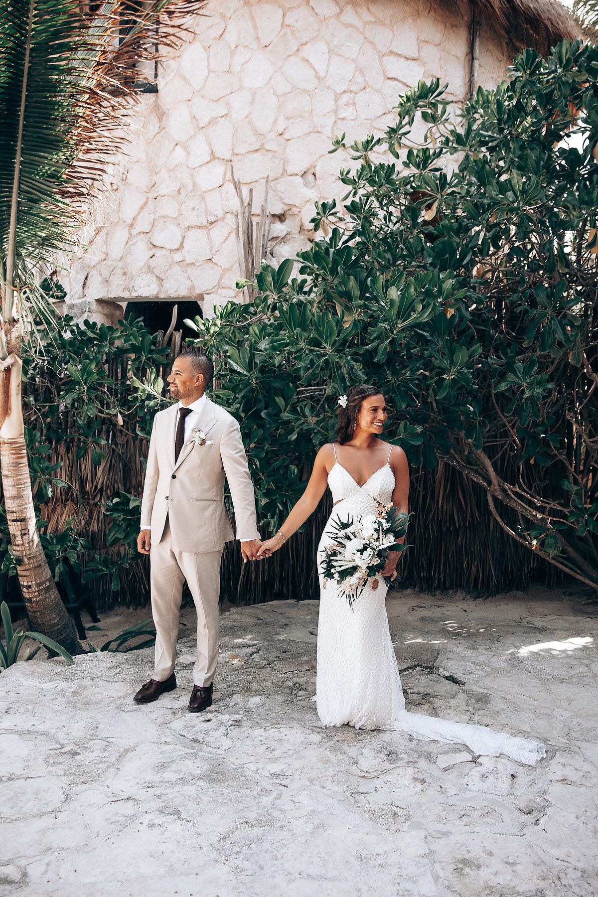 Wedding couple in white attire posing against a stone wall surrounded by tropical foliage.