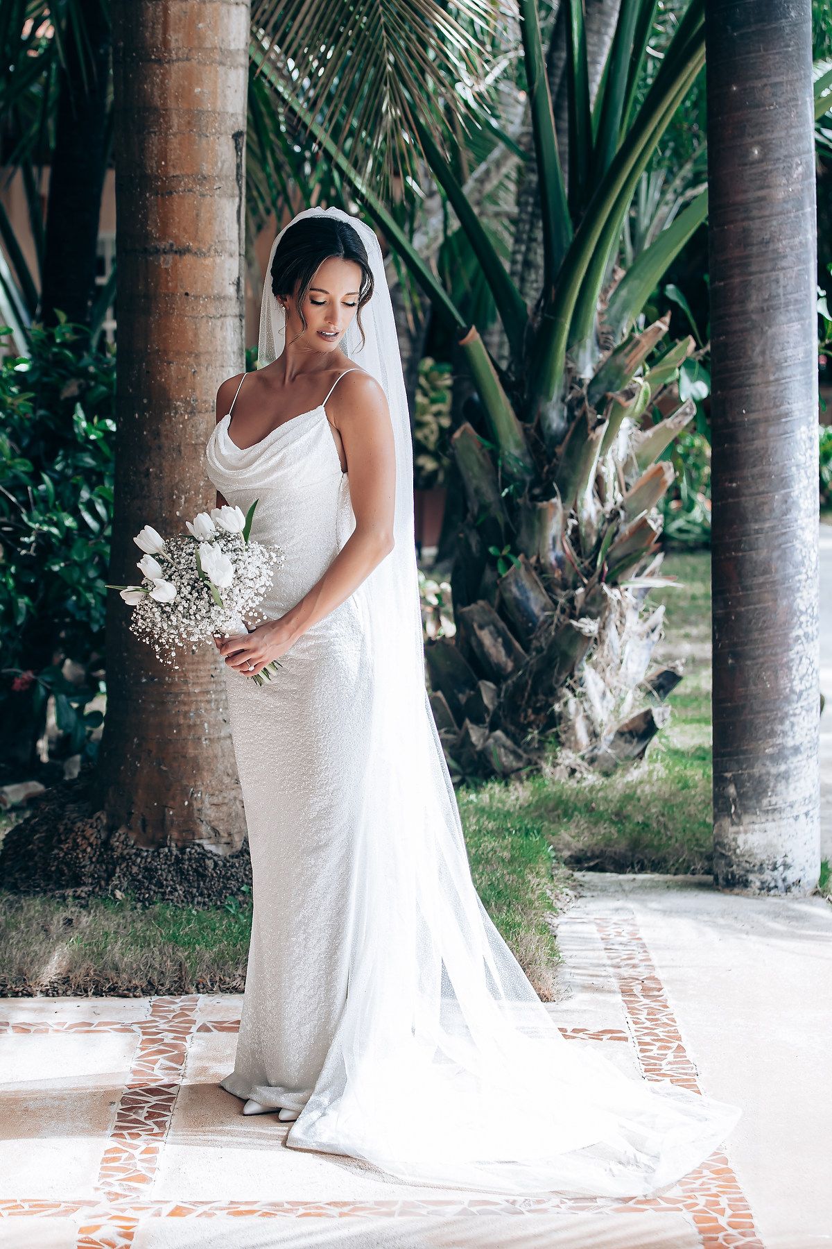Bride in sleek white dress holding bouquet while posing by palm trees.