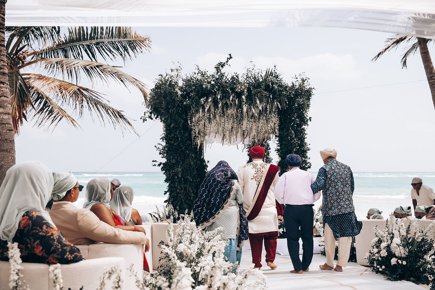 Beachfront wedding ceremony setup with floral arch and guests seated under palm trees.