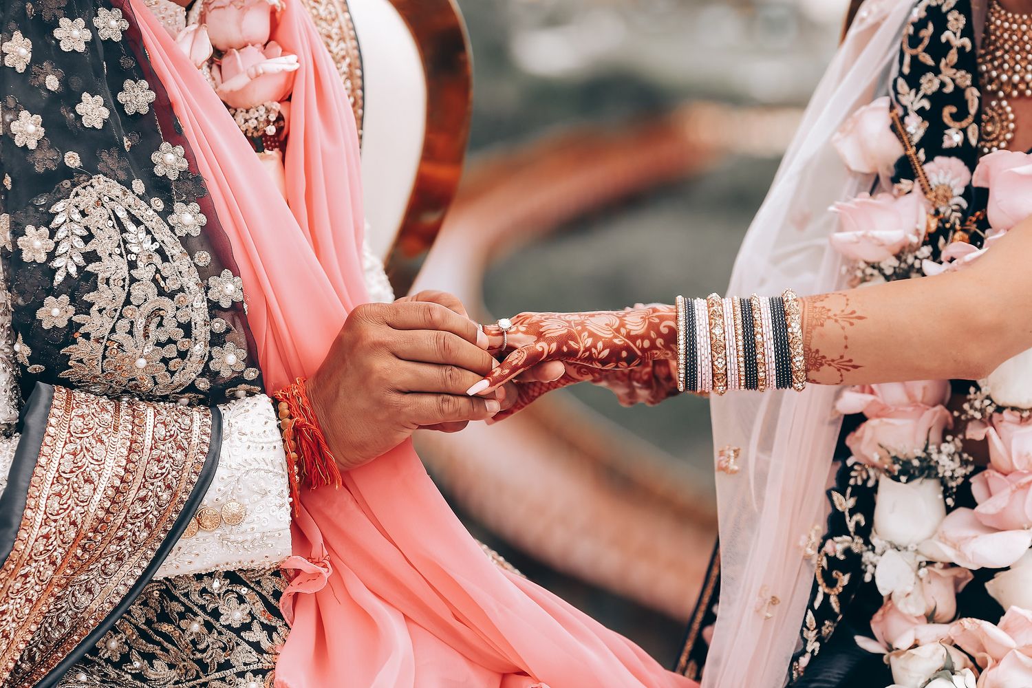 Close-up of hands adorned with traditional Indian wedding jewelry and henna exchanging rings during ceremony.