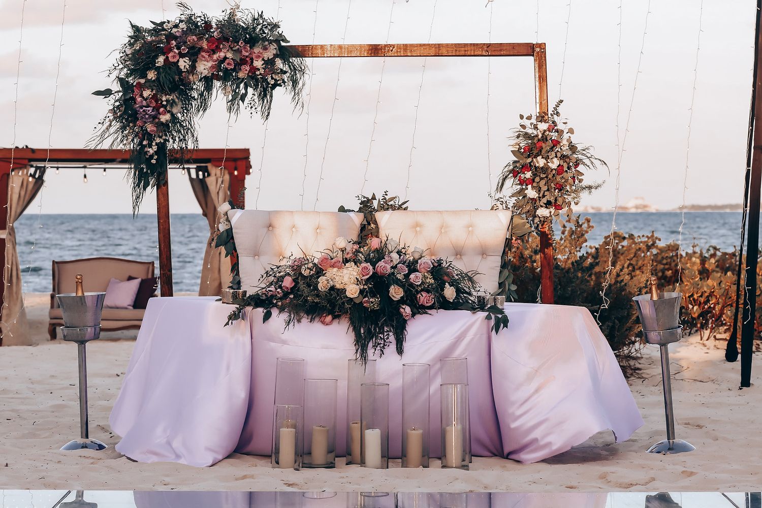 Romantic beach wedding setup with wooden arch decorated with pink and white flowers and flowy fabric at sunset.
