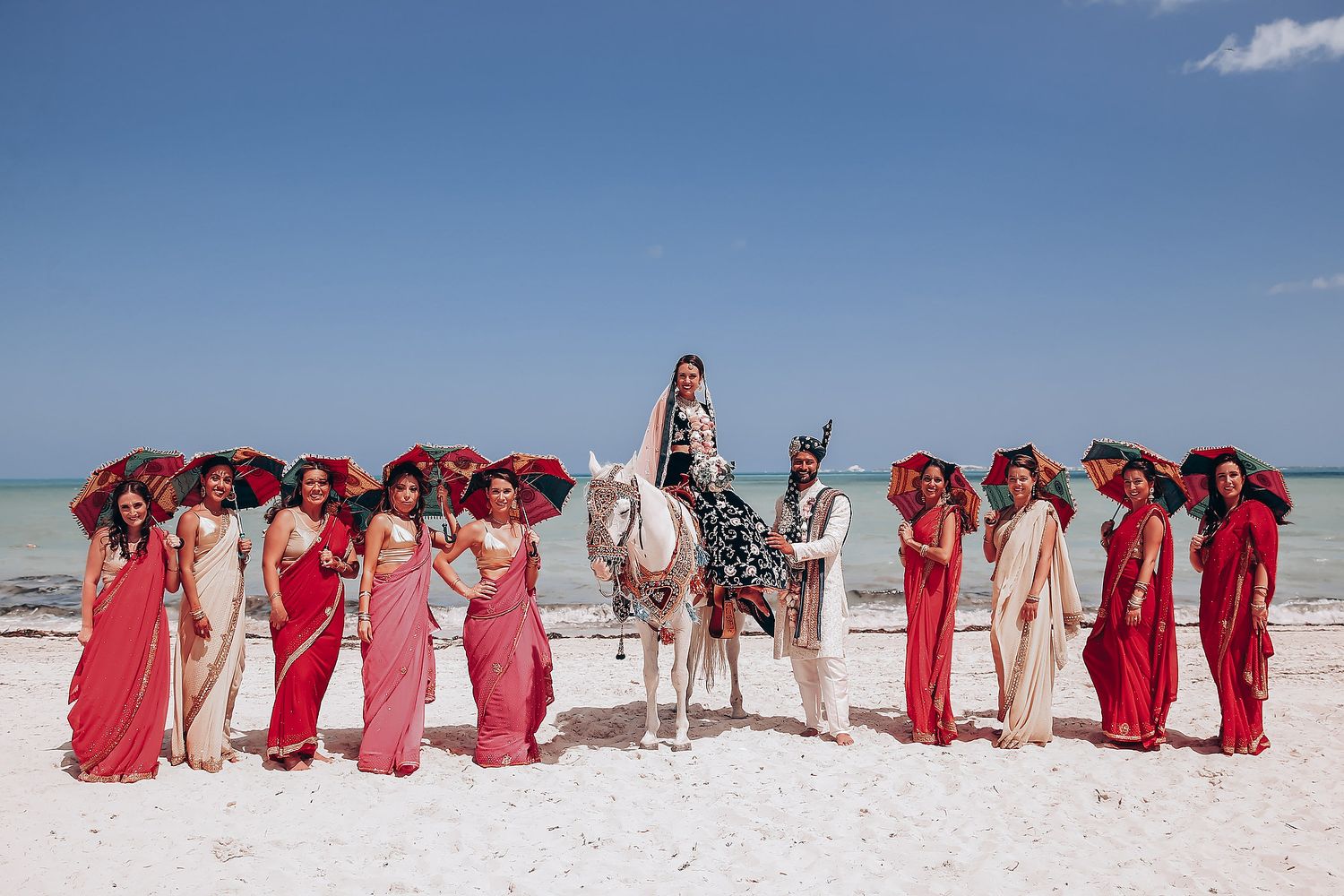 Wedding party in red dresses lined up on pristine white sand beach under blue sky.