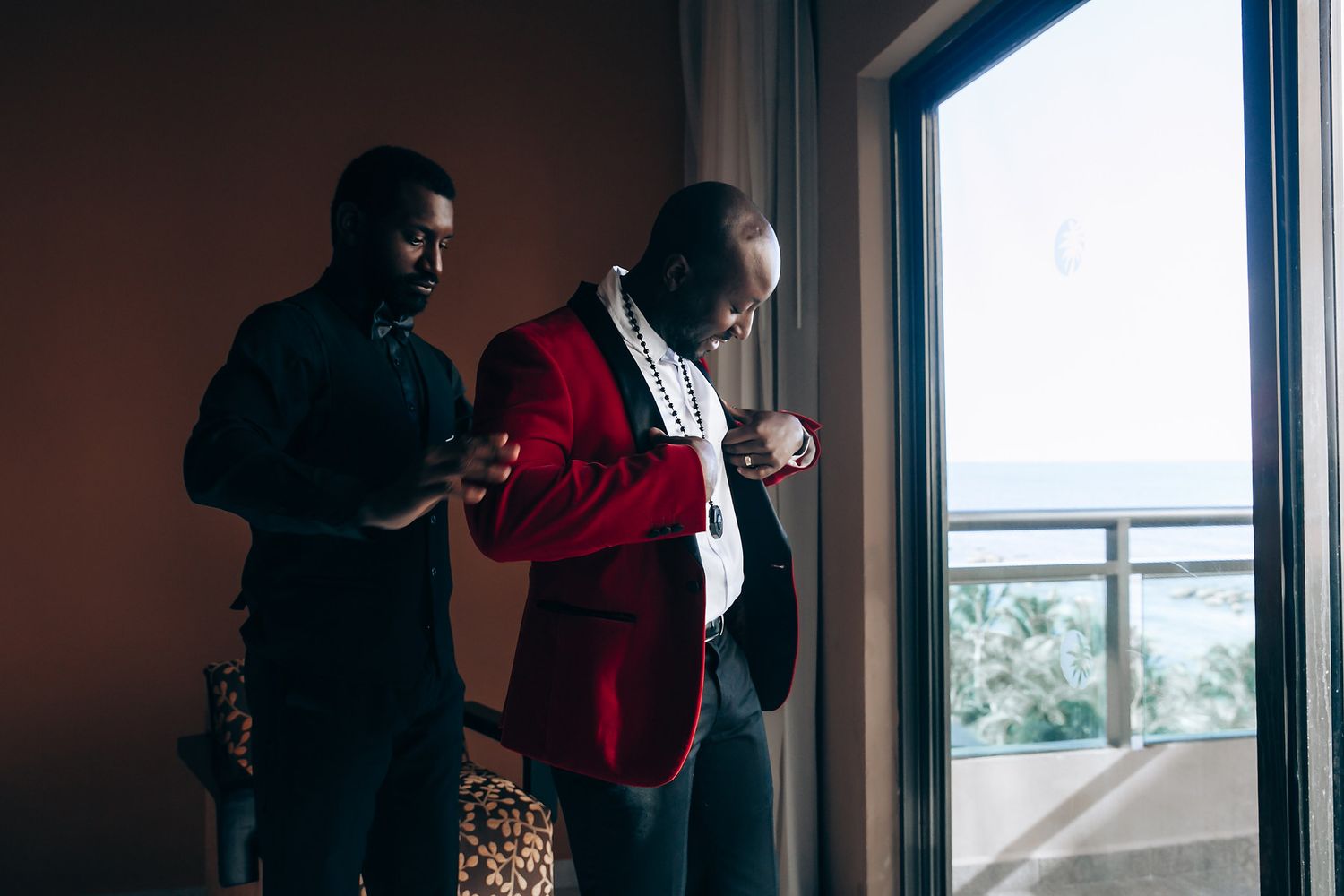 Two people in formal attire stand by a window, one wearing a striking red jacket.