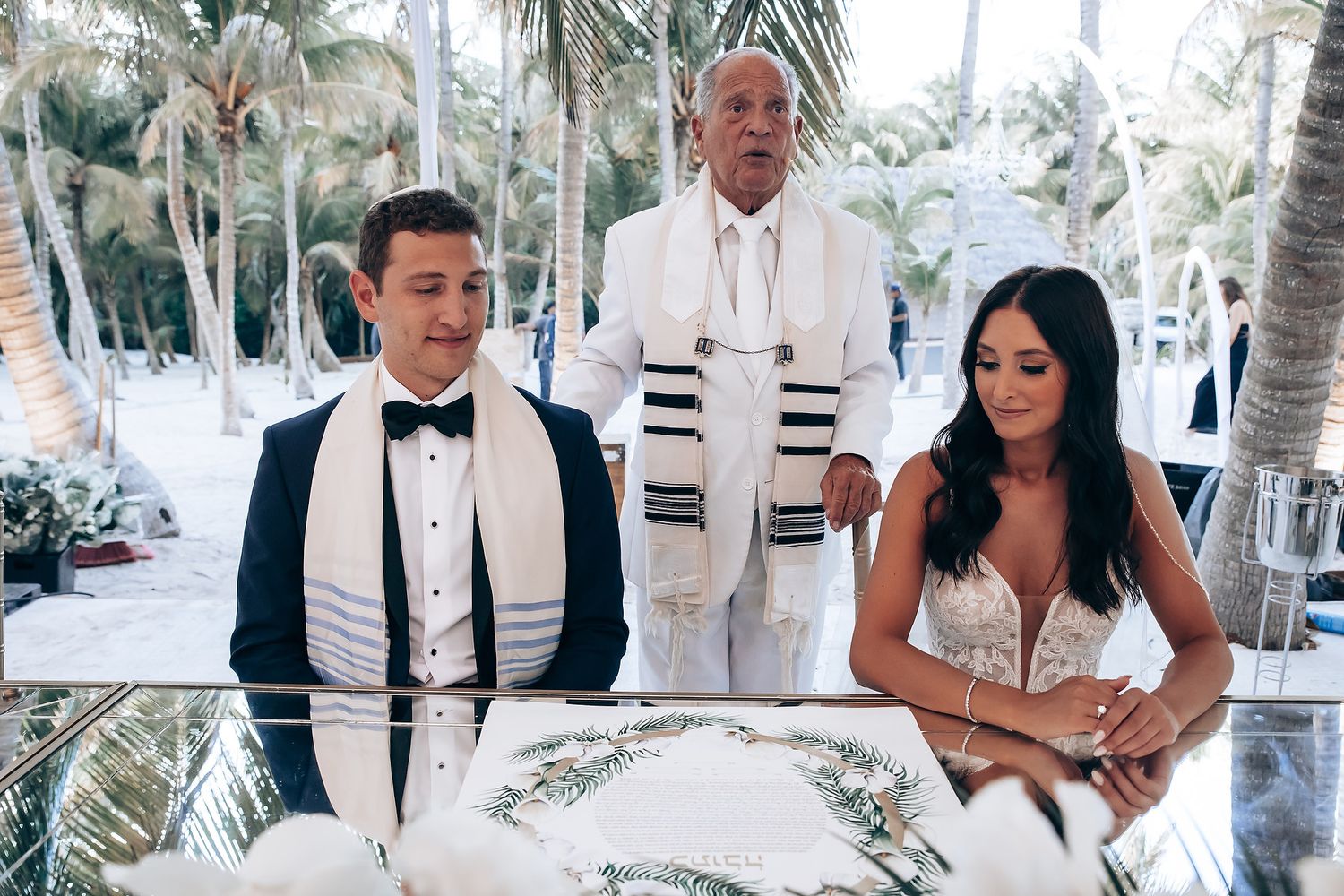 A romantic beach wedding ceremony with palm trees and white decor along a tropical shoreline.