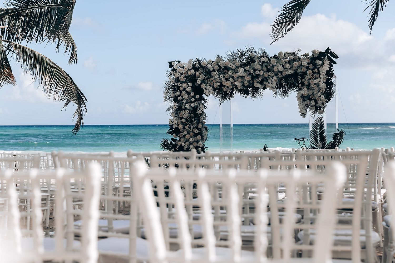 A beachfront wedding ceremony setup with white chairs and tropical palm trees overlooking turquoise ocean waters.