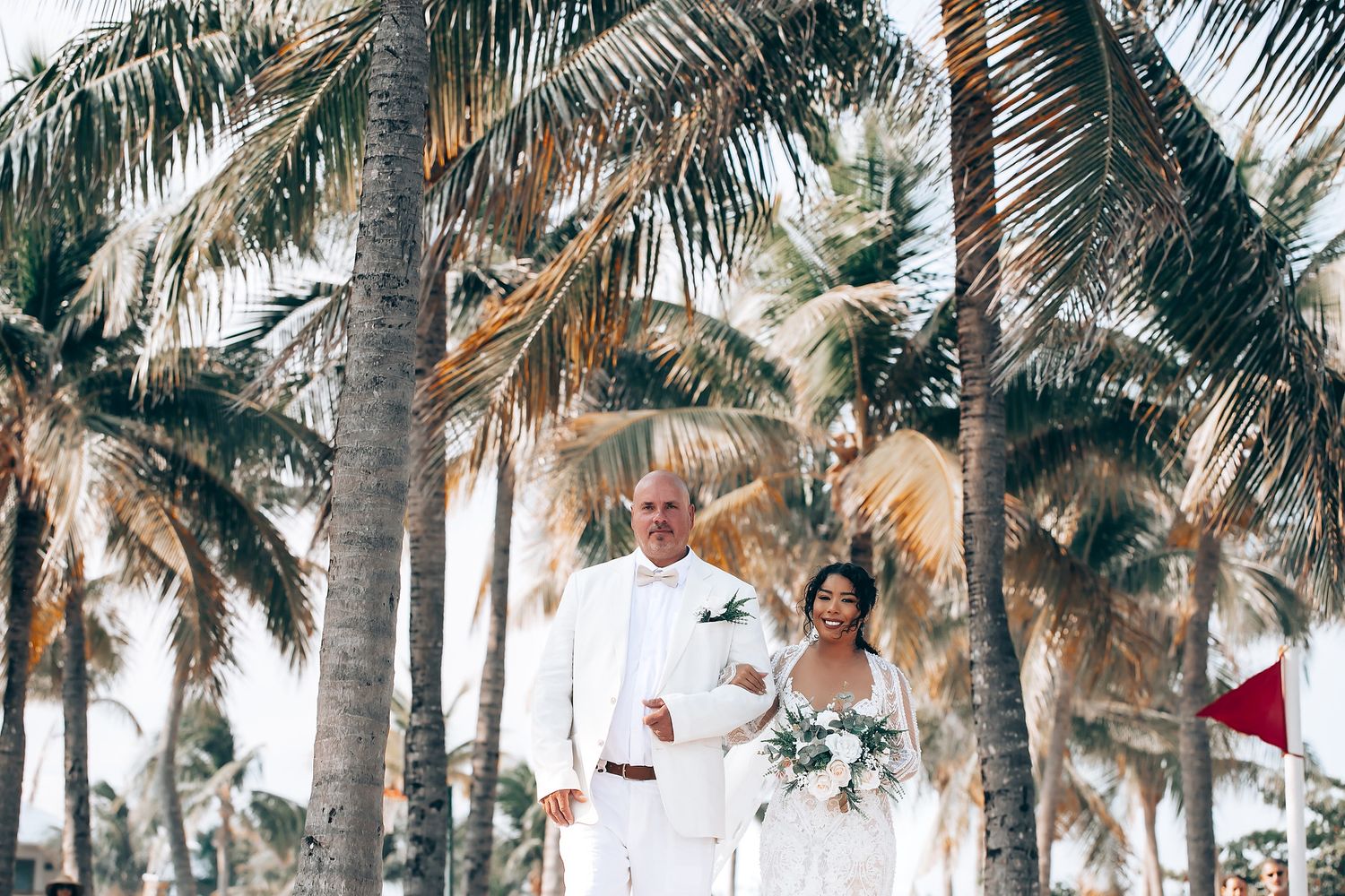 Palm trees line a tropical beach setting with guests in white attire gathered for a celebration.