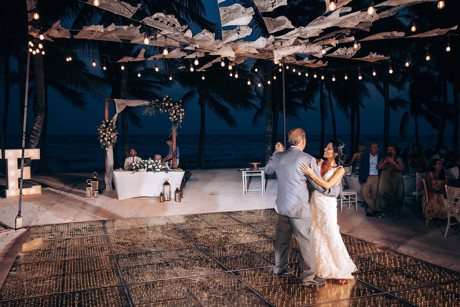 First dance at evening beach wedding reception under string lights and decorative ceiling.