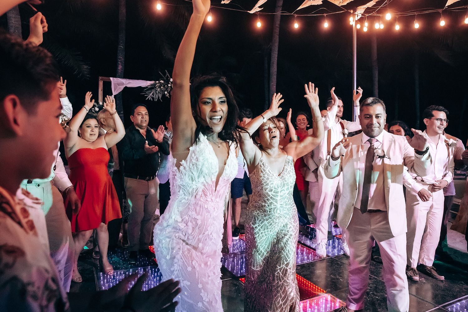 Wedding guests dance and celebrate with raised arms under string lights at a beach reception.