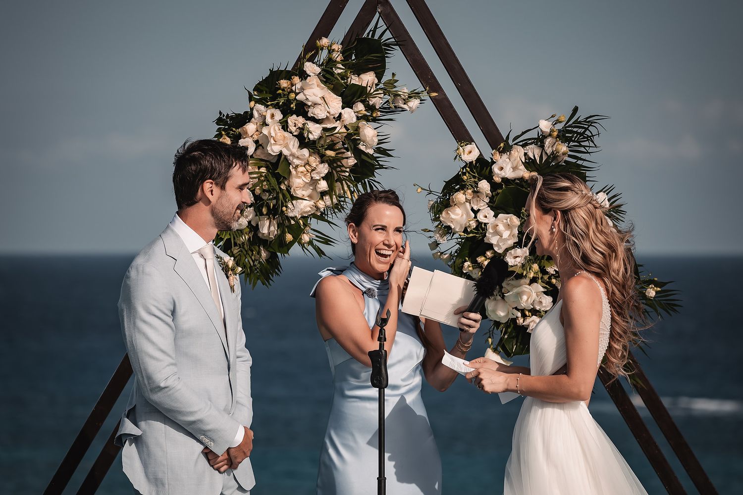 Seaside wedding ceremony with modern triangular arch and floral arrangements.