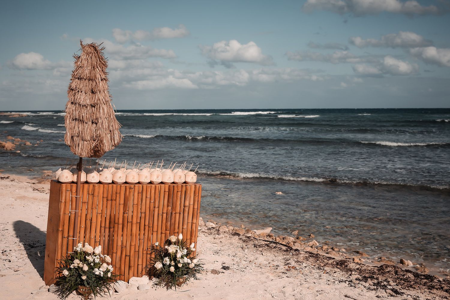 Rustic wooden beach wedding ceremony setup with floral arrangements and ocean waves in the background.