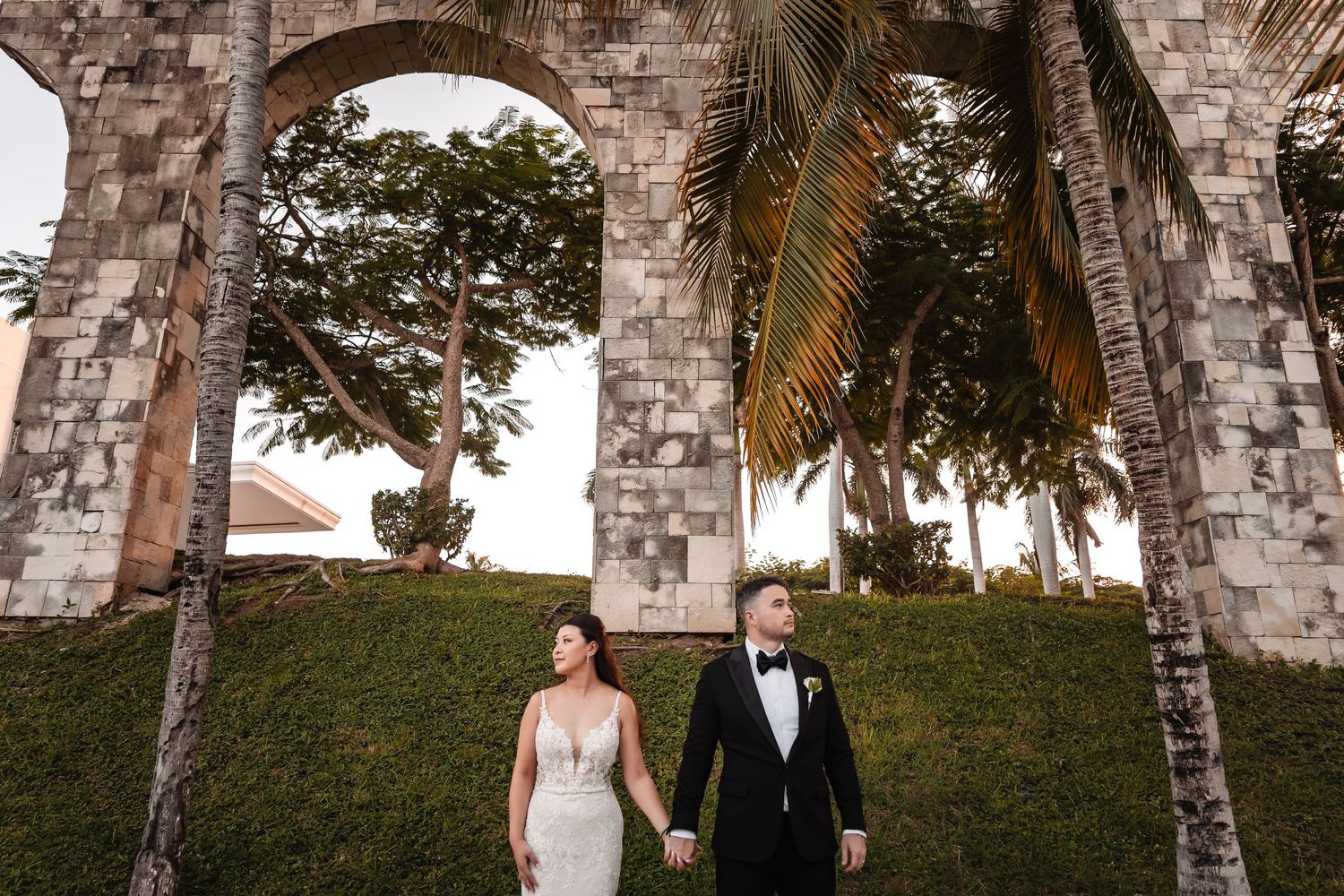 Couple walks beneath stone archway with palm trees and grass hill in background.