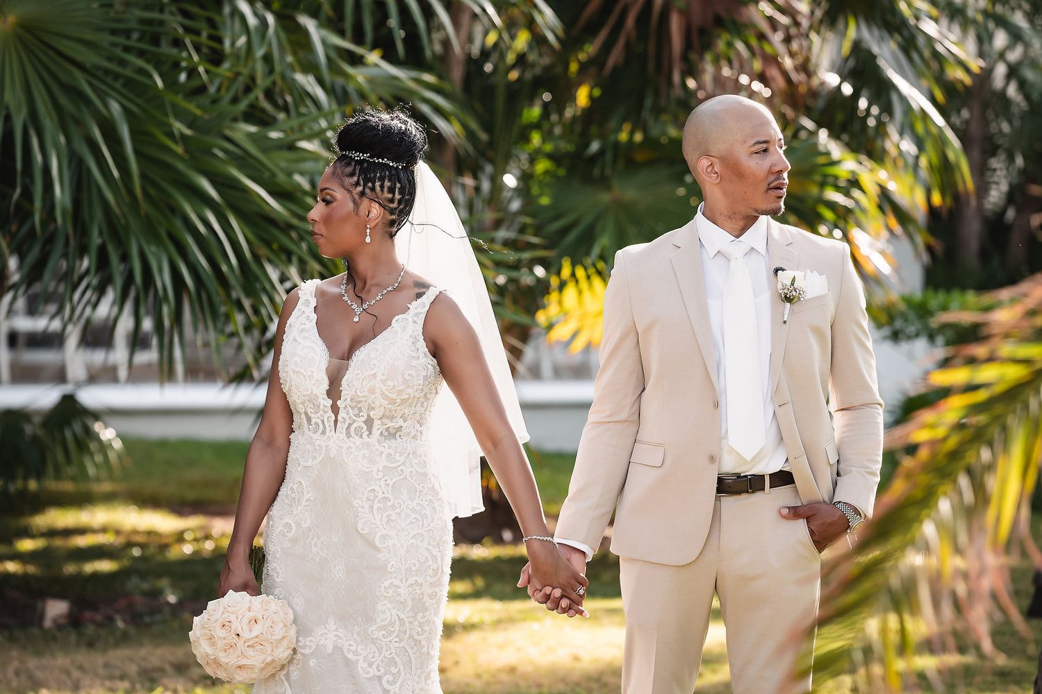 A couple in elegant wedding attire poses together in a lush tropical garden setting.