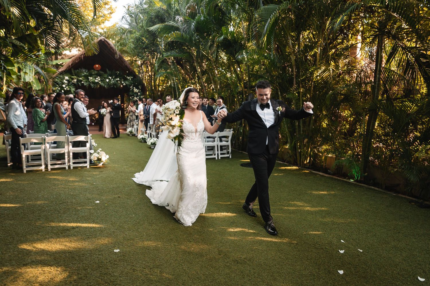 Wedding ceremony guests seated in white chairs on a manicured lawn with tropical landscaping.