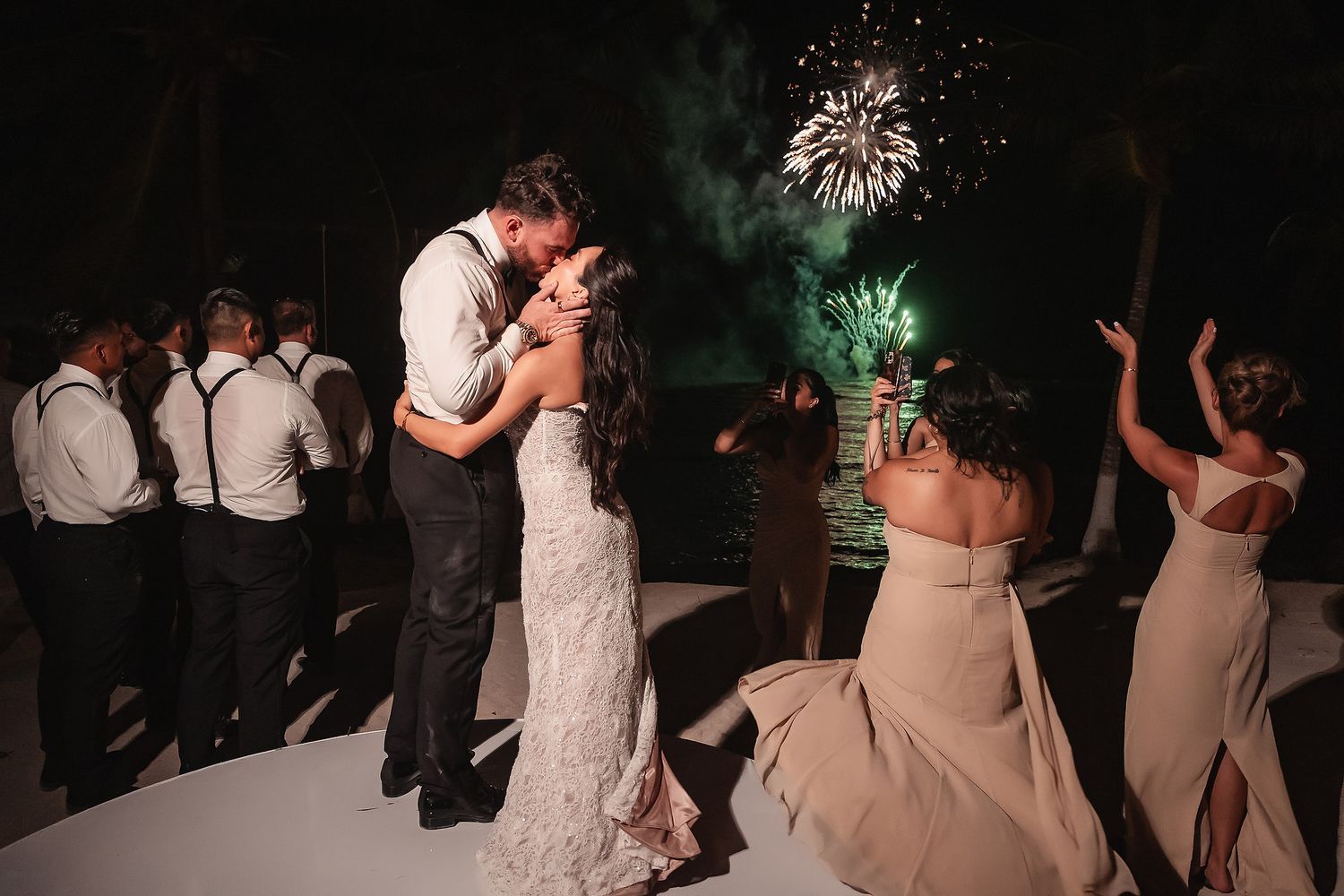 Wedding guests celebrate on a beach with colorful fireworks lighting up the night sky.