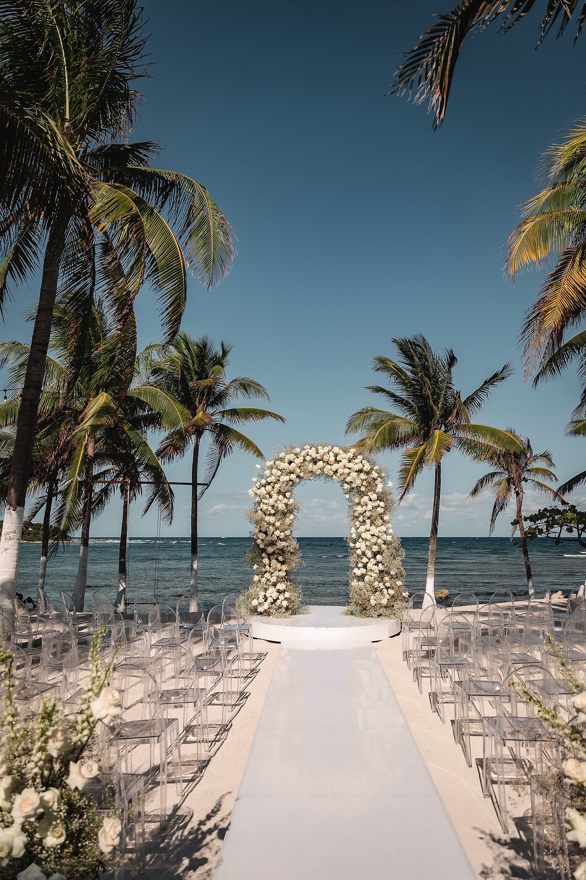 Beachfront wedding ceremony setup with white chairs and floral arch overlooking the ocean and palm trees.