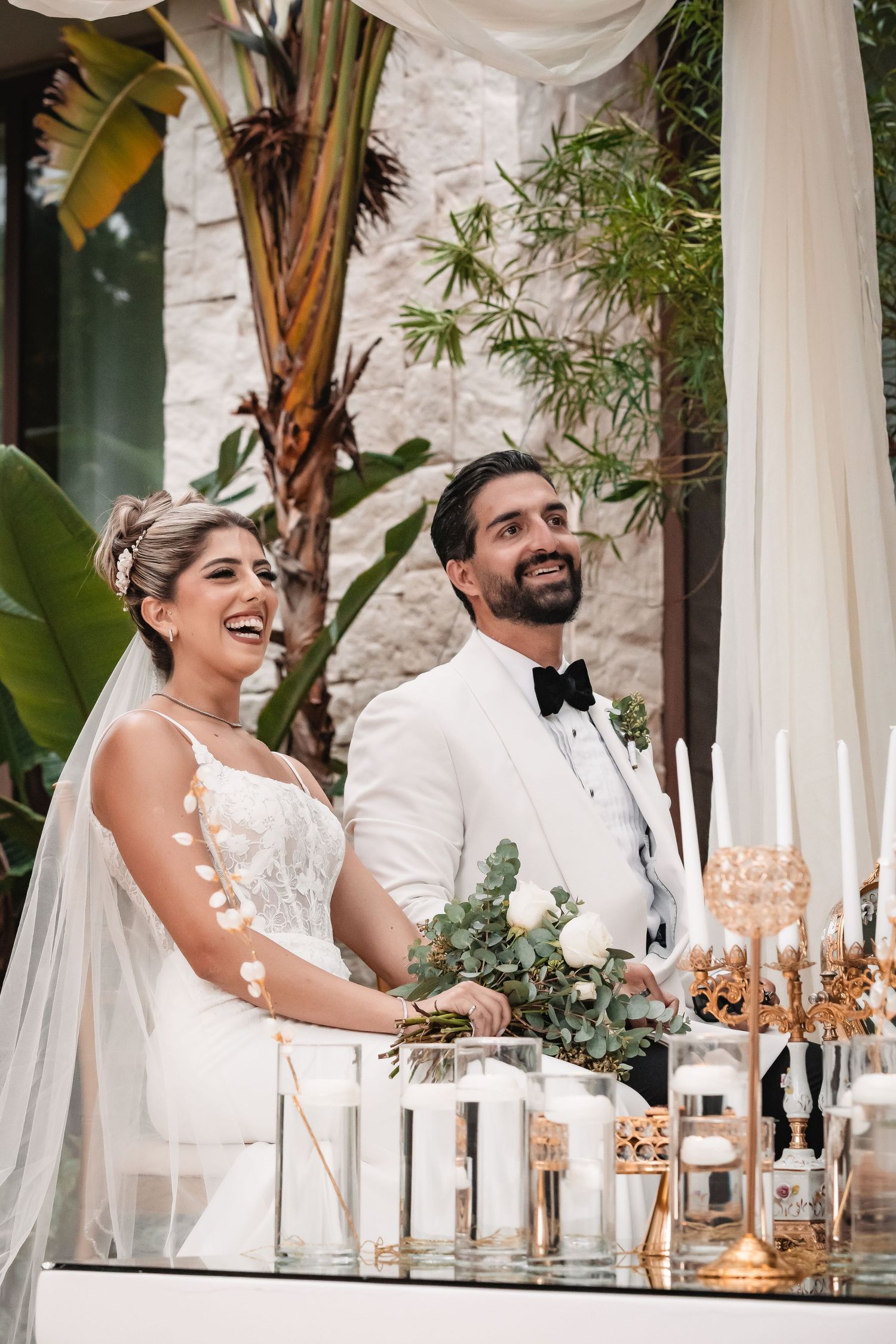 Wedding couple stands at reception table with elegant candles and greenery decor.