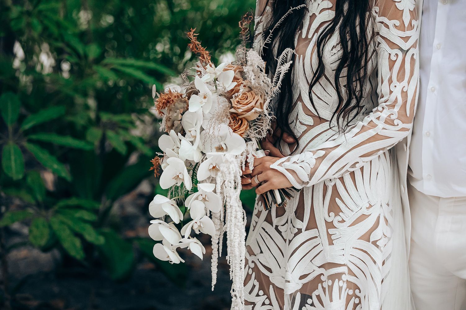 A bride in a white lace wedding dress holds a bouquet of white flowers against a natural outdoor backdrop.