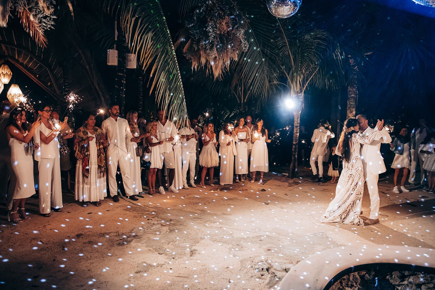 Wedding guests dance and celebrate on a confetti-covered dance floor at an outdoor evening reception.