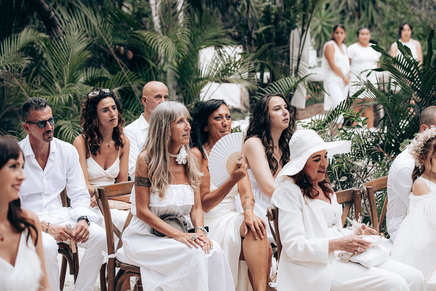 Wedding guests dressed in white gather outdoors for a celebration surrounded by tropical greenery.