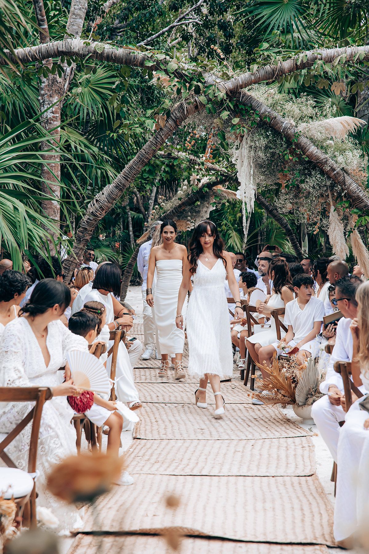 A group of people dressed in white gather for a celebration under palm trees on a tropical patio.