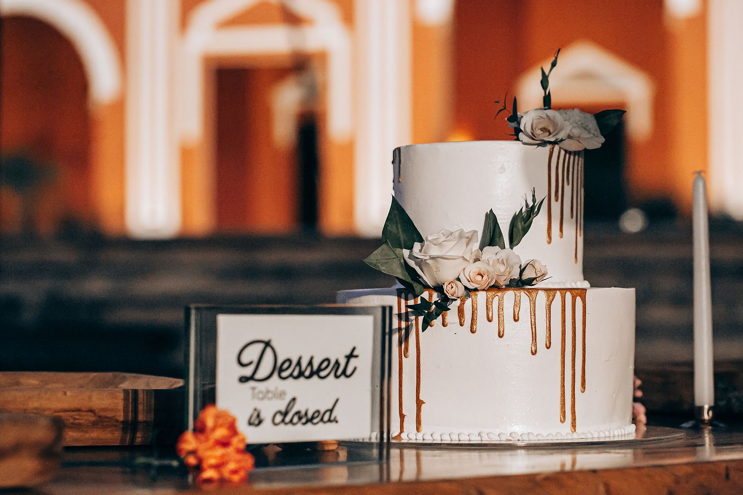 Modern white wedding cake with chocolate drip details and fresh flowers displayed on a rustic wooden table.
