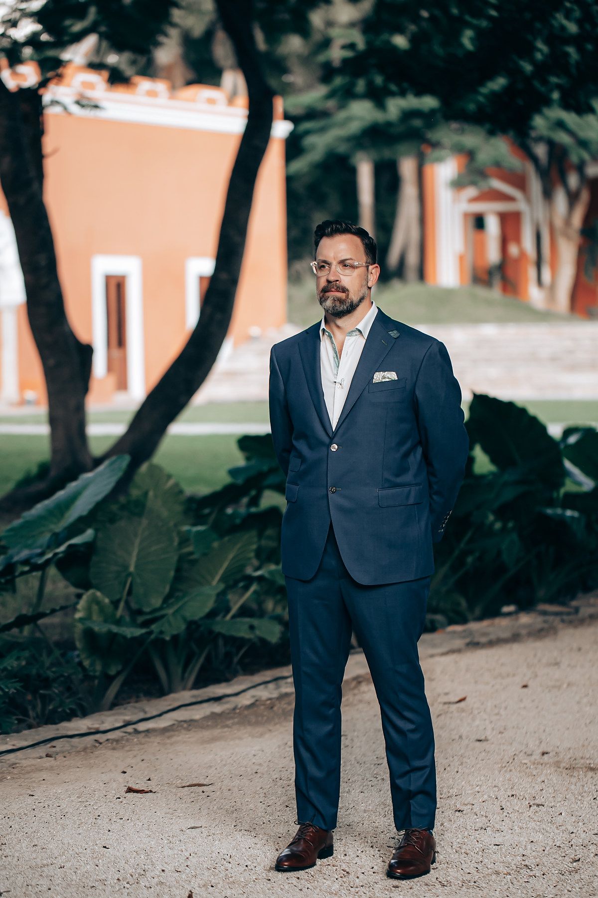 A person in a navy blue suit stands outdoors on a path near trees in a professional portrait setting.