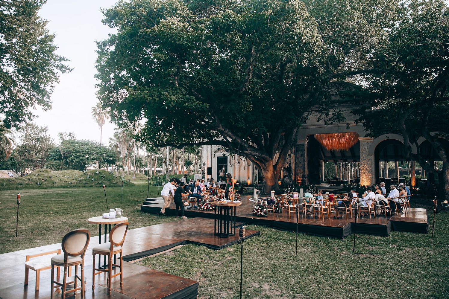 A rustic outdoor wedding venue features wooden chairs arranged in rows beneath towering trees.
