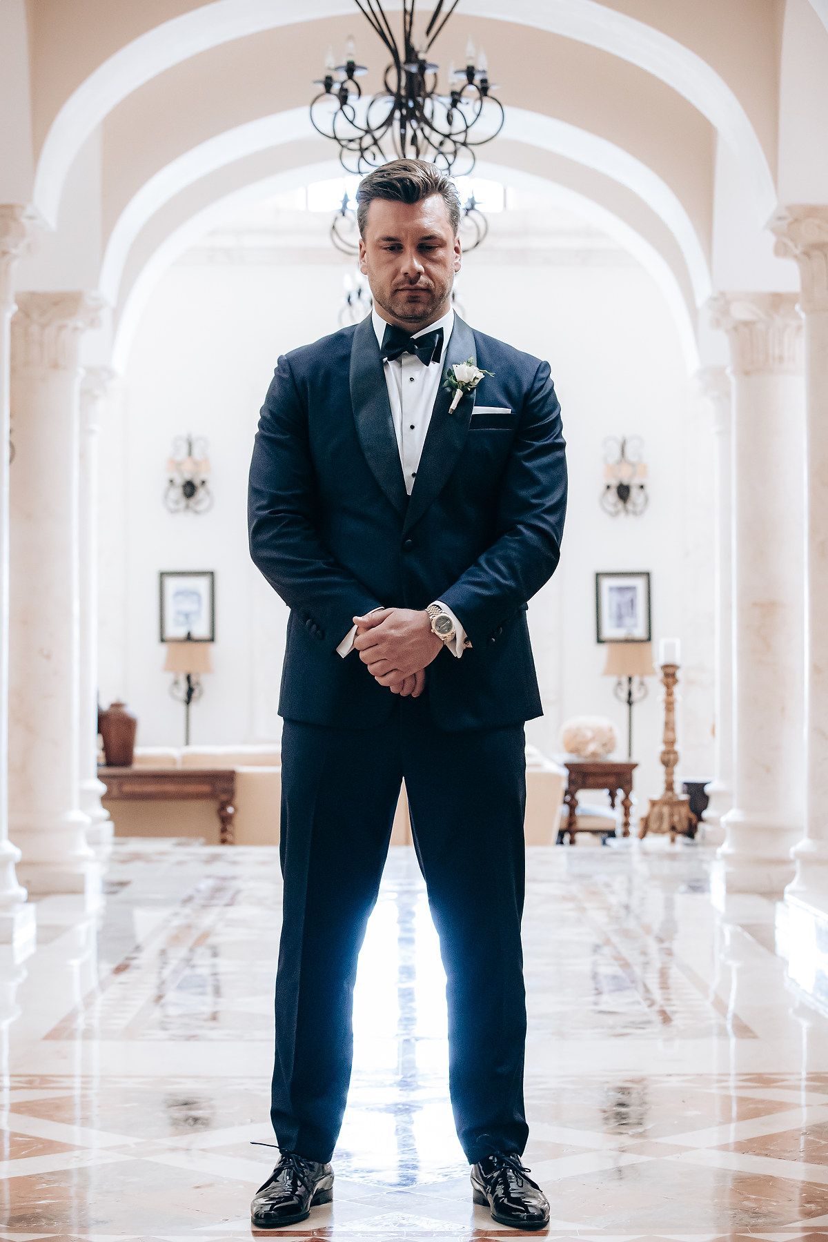 A groom in a black tuxedo poses in an elegant white marble hallway with arched columns and chandeliers.
