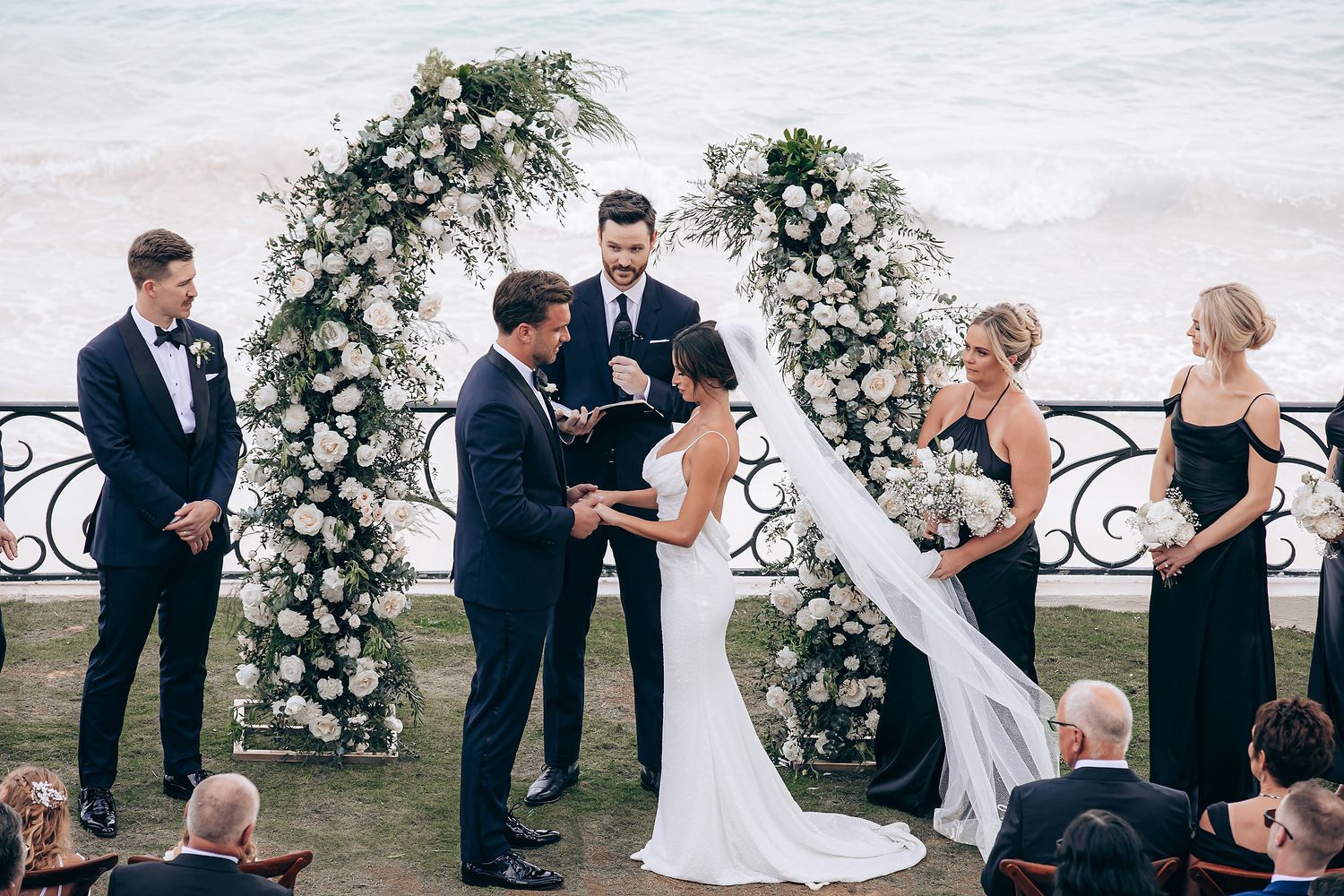 Wedding party poses for photos on balcony with white floral arrangements and seaside view.