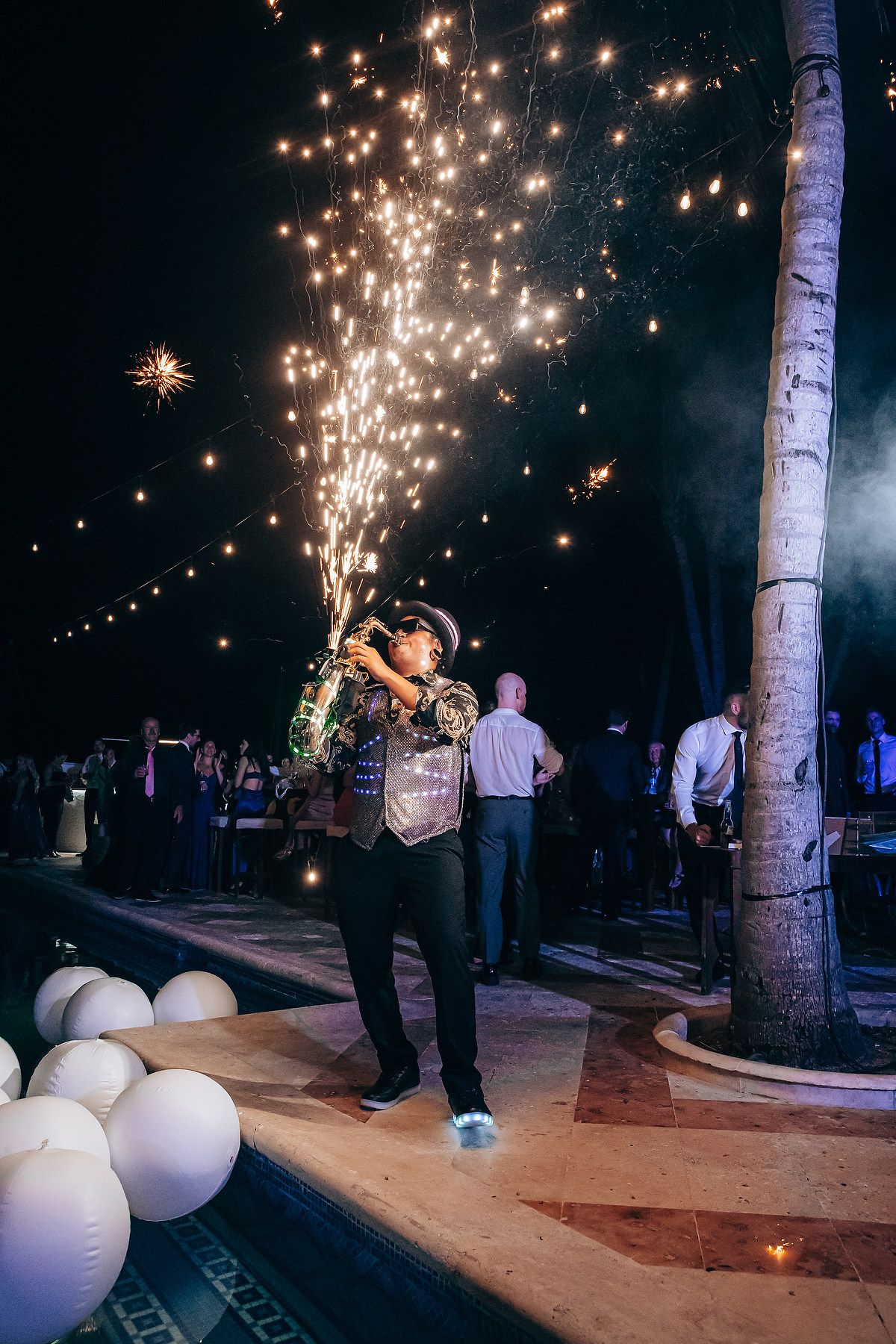A person holds a sparkling firework at an outdoor nighttime celebration with white balloons and palm trees.