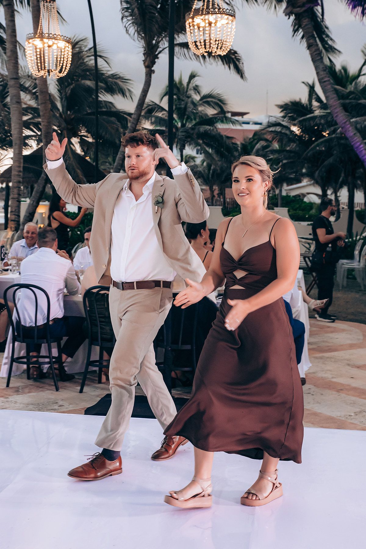Couple dancing together at a tropical beach wedding reception with palm trees and chandeliers in the background.
