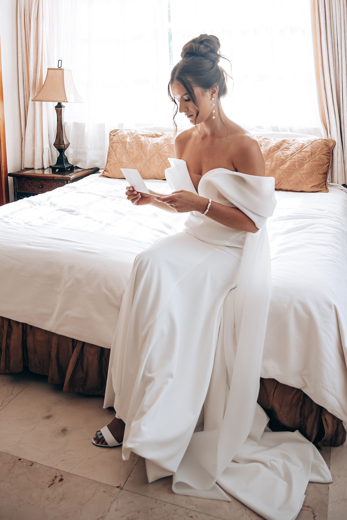 A woman in an elegant white off-shoulder gown sits on a bed in a sunlit room with beige decor.