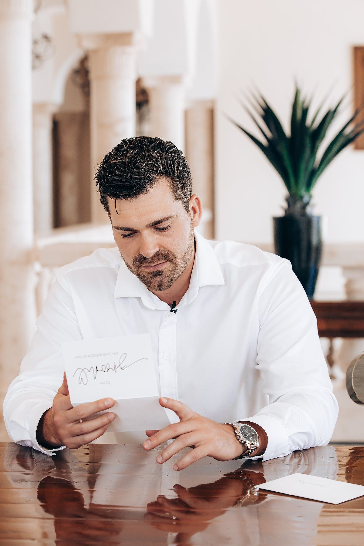 Person in white dress shirt reading documents at wooden desk with houseplant in background.