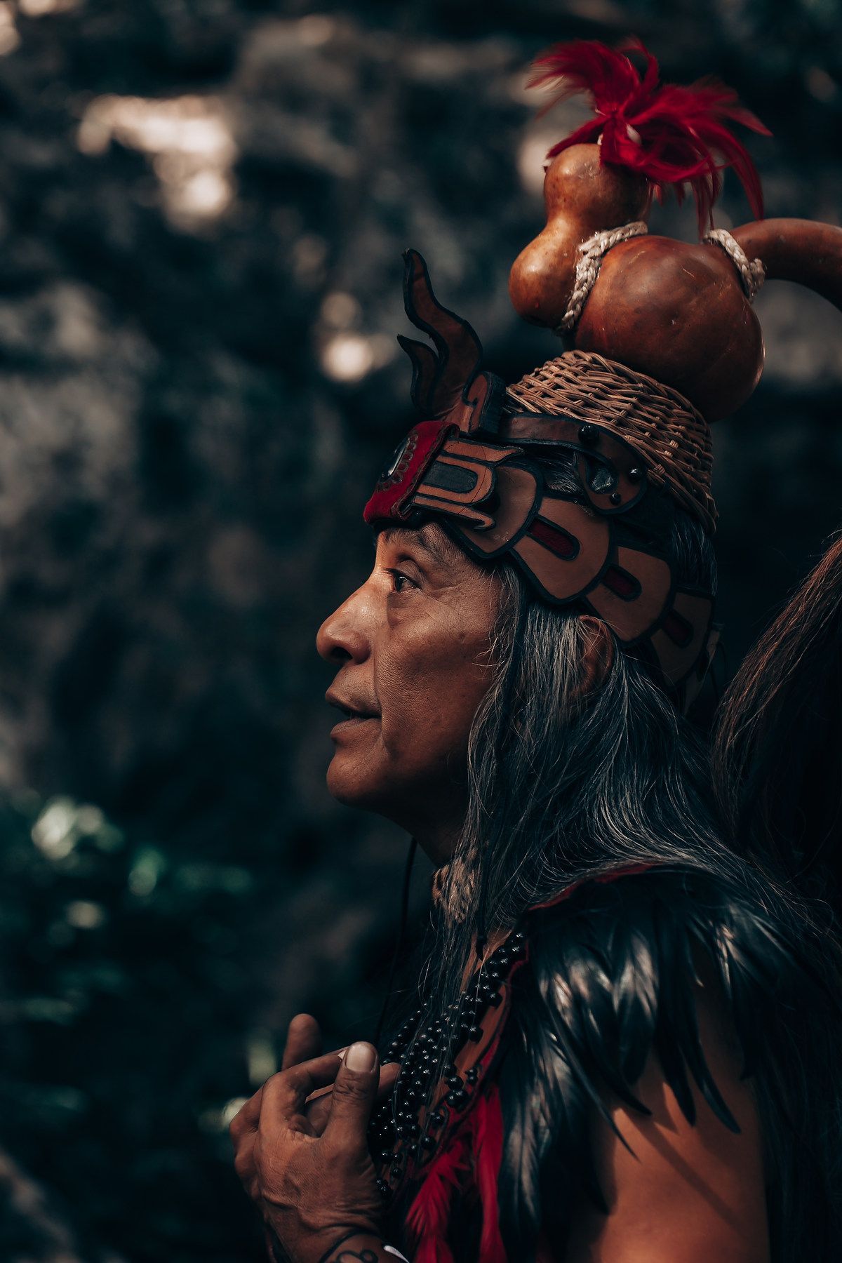 Traditional Mayan healer wearing ceremonial headdress with feathers and red decorations in natural setting.