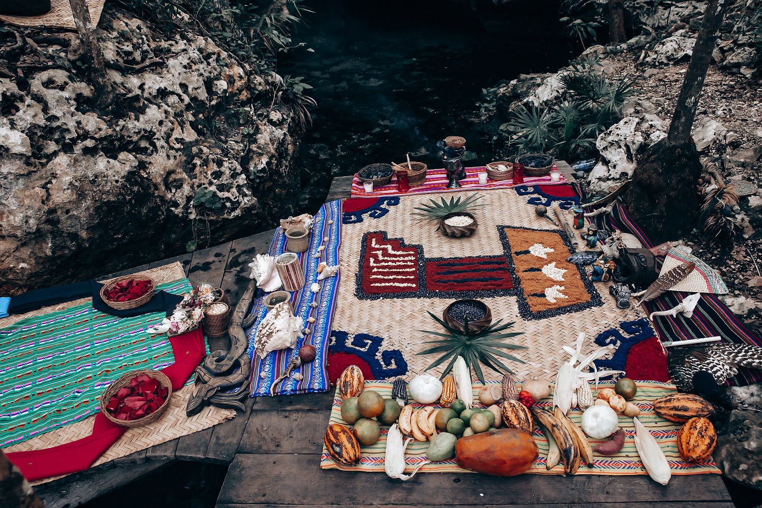 Colorful bohemian picnic setup with rugs and cushions arranged on wooden platform in a cave setting.