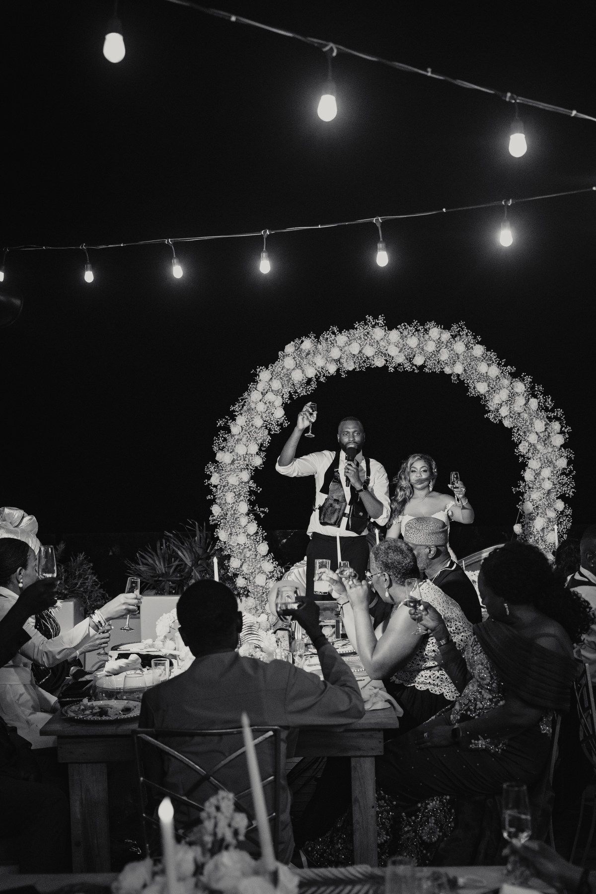 Black and white photo of wedding toast under string lights with floral arch backdrop at night.
