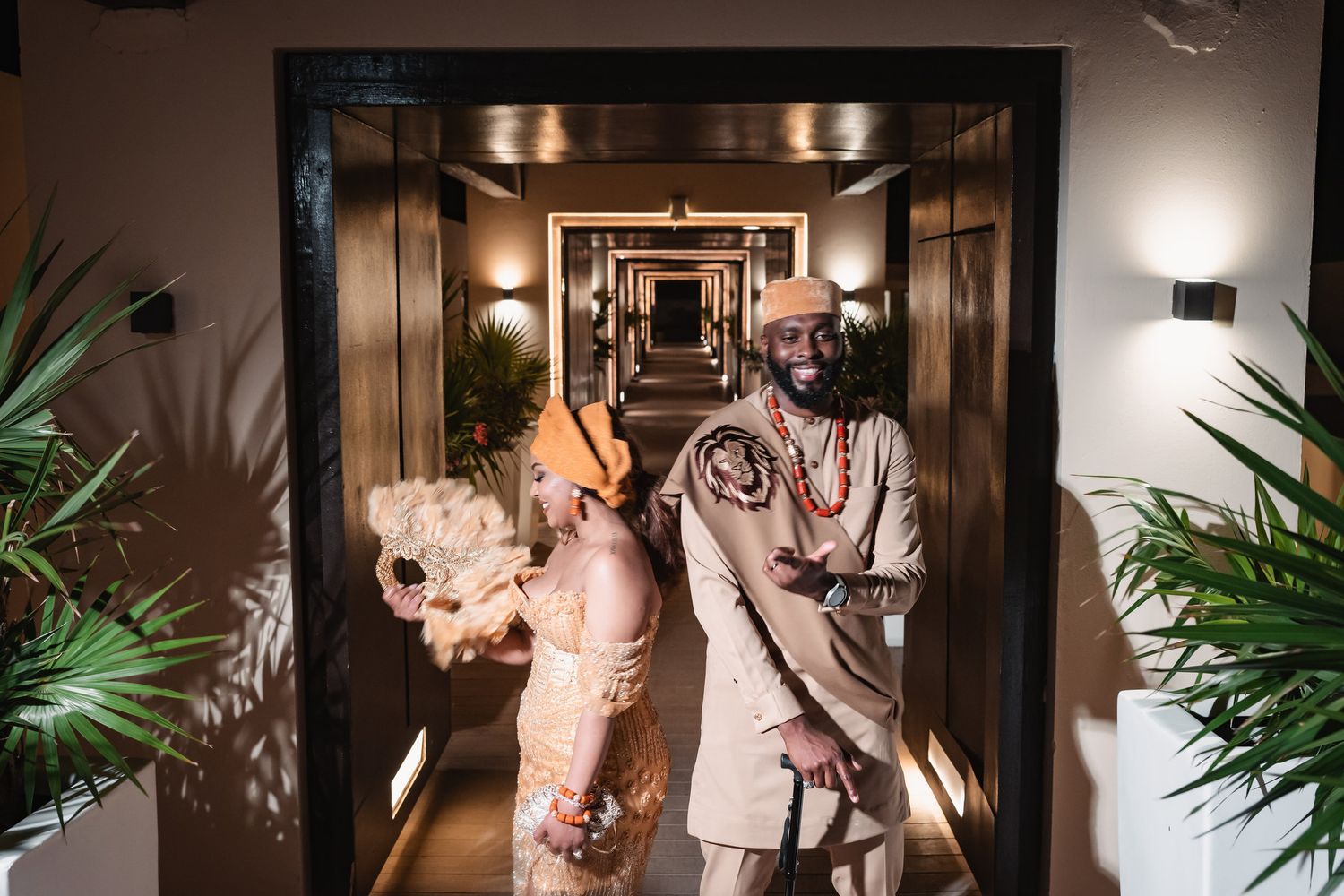 Couple in traditional Nigerian wedding attire walk elegantly through a warmly lit hotel corridor.