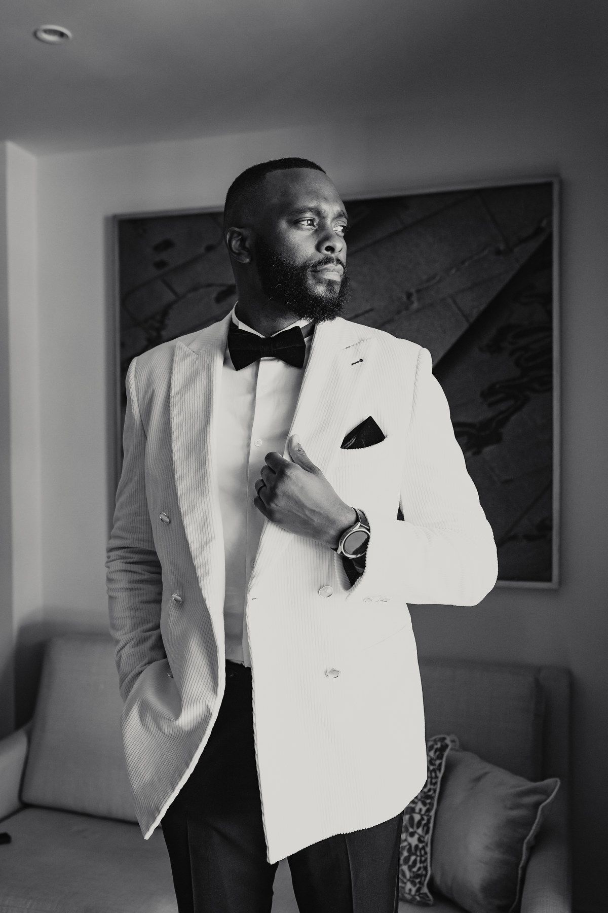 Elegant groom in white tuxedo jacket and black bow tie posing in black and white portrait.