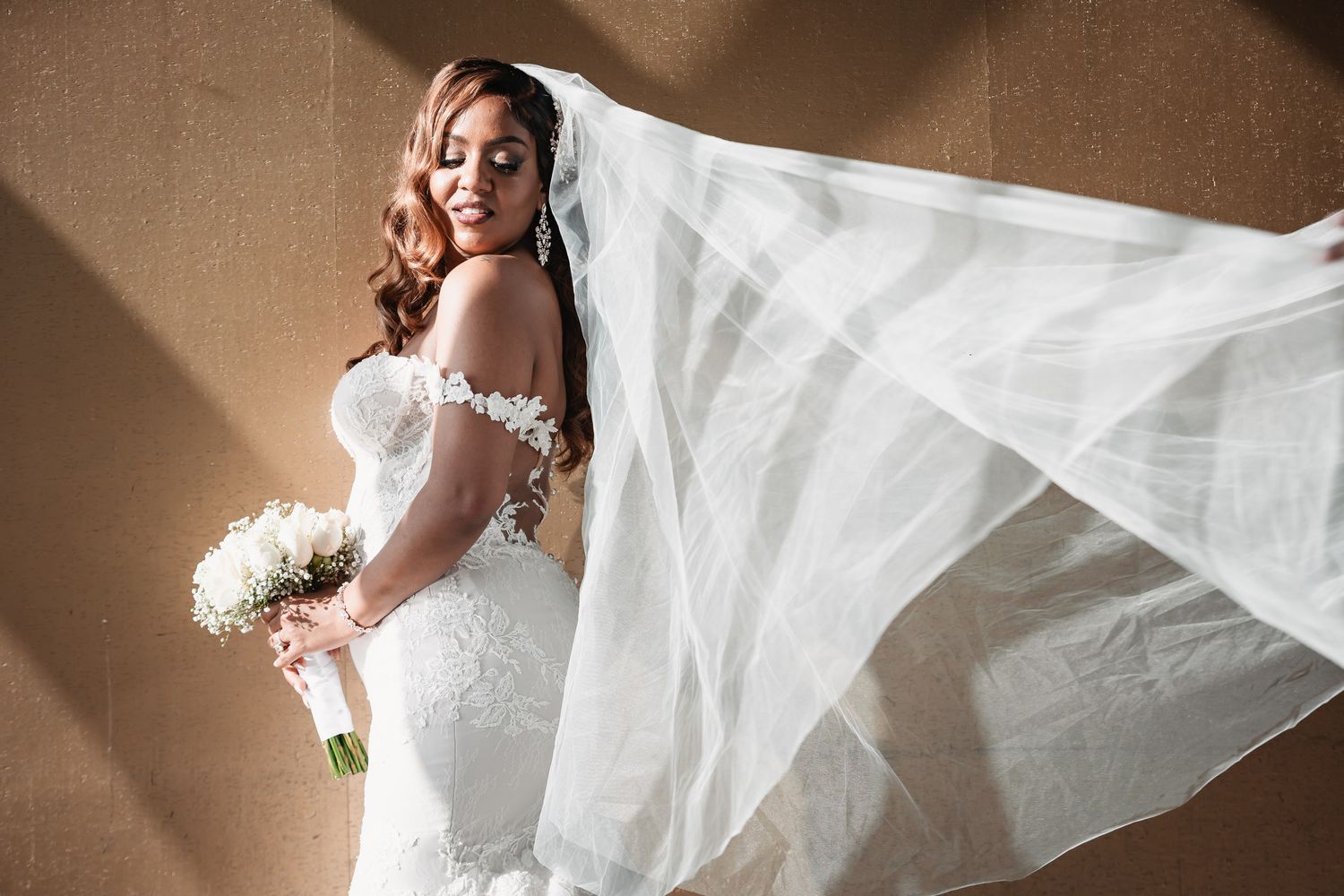 Elegant bride in lace wedding gown with flowing veil holding white bouquet in soft natural light.