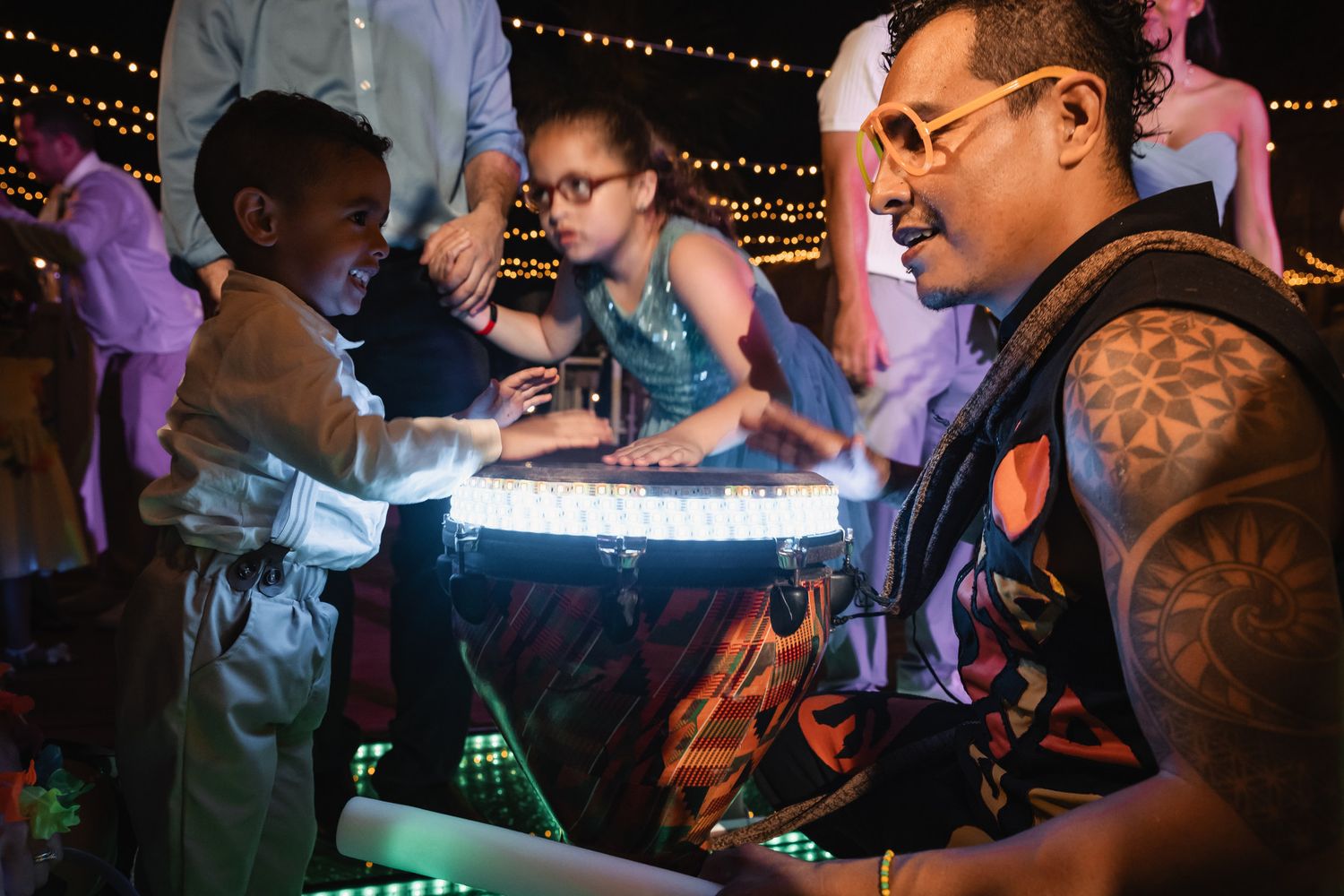 A tattooed DJ entertains a young child at a glowing DJ booth during a lively evening event.