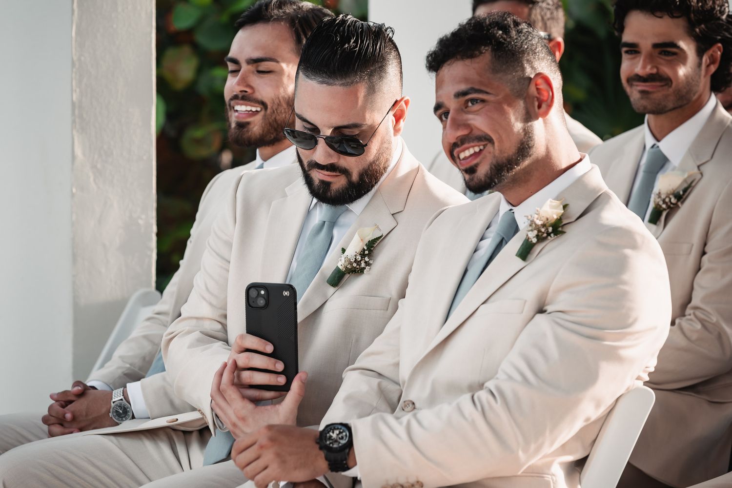 Groomsmen in cream suits smile and take photos on phones during wedding ceremony.