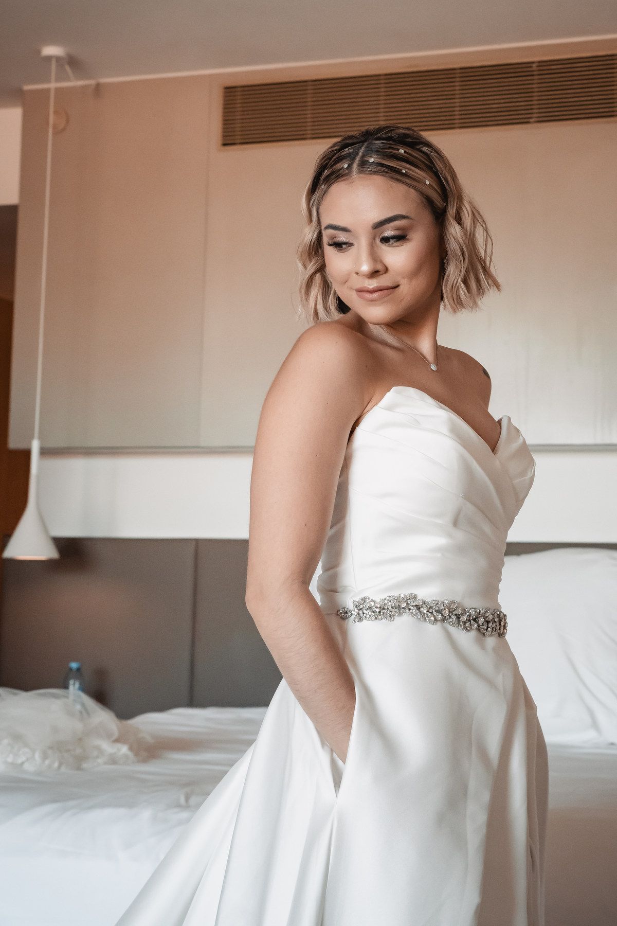 Elegant bride in strapless white satin wedding gown with crystal belt posing in hotel room.